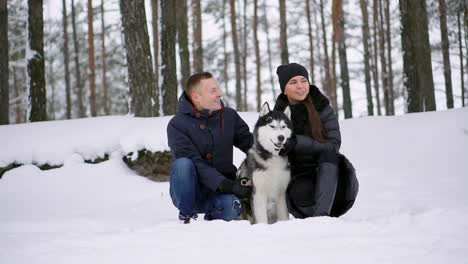 A-man-and-a-woman-sitting-hugging-a-dog-Siberian-husky-in-the-winter-forest-smiling-and-looking-at-each-other-and-at-the-camera.-Slow-motion-happy-family